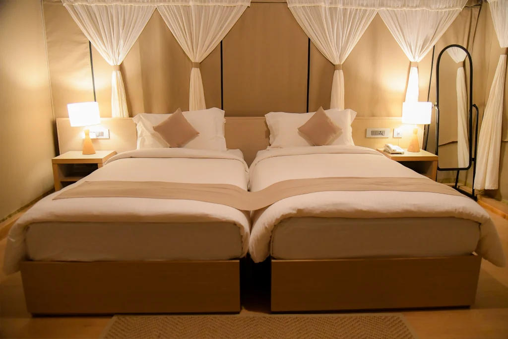 Two neatly made twin beds with white linens and beige accents stand side by side in a softly lit room. Each bed has a beige pillow, and there are bedside tables with lamps on both sides, with sheer curtains in the background.