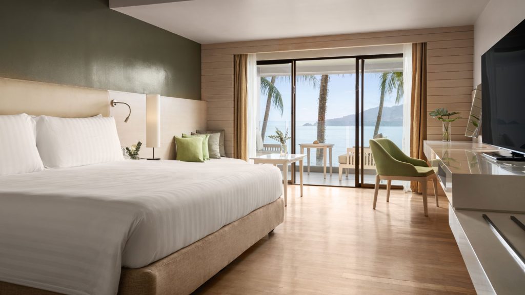 A modern hotel room with a large bed, green accent chair, and a balcony overlooking the ocean with palm trees. Sunlight brightens the room through sliding glass doors.