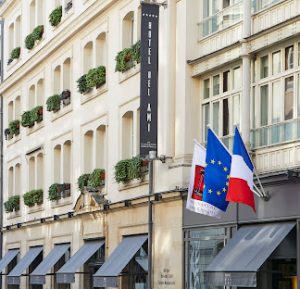 A street view of Hôtel Bel Ami with flower boxes on windows, a vertical hotel sign, and three flags—European Union, France, and a red-and-black flag—displayed at the entrance.