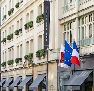 A street view of Hôtel Bel Ami with flower boxes on windows, a vertical hotel sign, and three flags—European Union, France, and a red-and-black flag—displayed at the entrance.