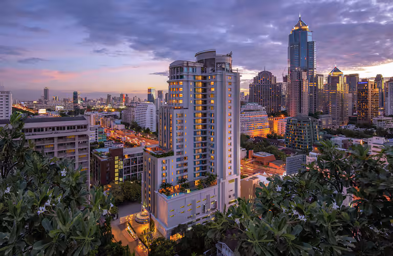 A cityscape at sunset shows modern high-rise buildings with lights on, set against a dramatic, cloudy sky. Trees and greenery frame the foreground, blending urban and natural elements.