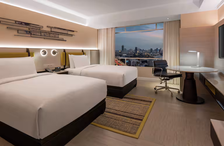 Modern hotel room with two double beds, a large window with a city skyline view, a desk with a chair, wall art above the beds, soft lighting, and neutral-toned decor.