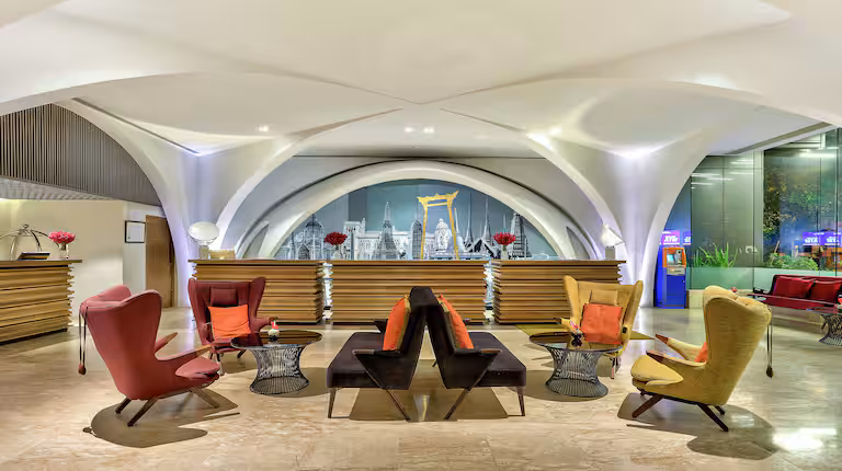 Modern hotel lobby with colorful armchairs and sofas arranged around small tables. Behind the seating area is a reception desk, and a large cityscape mural is displayed on the back wall. The space is brightly lit.