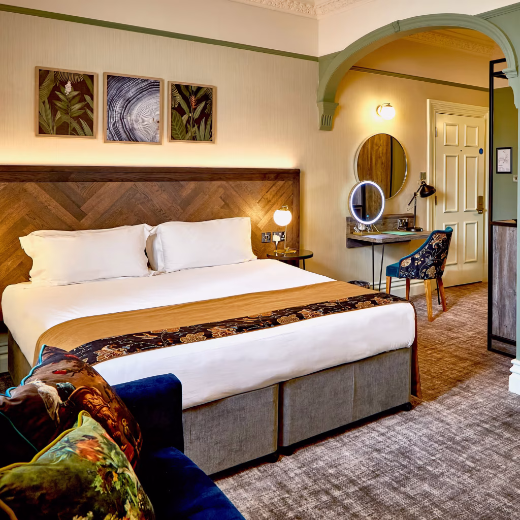 A stylish hotel room with a large bed, white bedding, a patterned throw, and headboard art. Theres a vanity with a mirror, a blue chair, and a couch with decorative pillows. Warm lighting creates a cozy atmosphere.