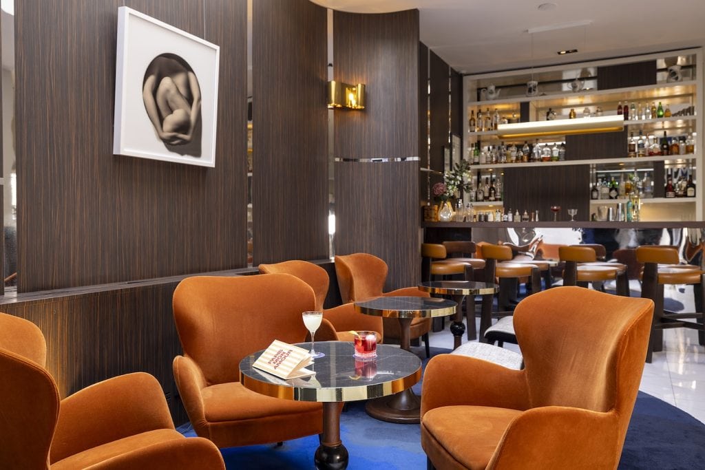 A stylish lounge bar with plush orange armchairs, round black tables holding drinks, and a modern bar in the background. Dark wood paneling and abstract wall art create a contemporary, inviting atmosphere.