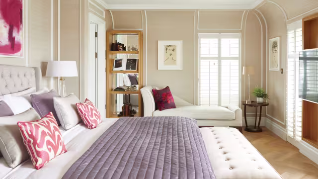 A stylish bedroom with a tufted bed, lavender quilt, patterned pillows, a bench, a chaise lounge by windows with shutters, wall art, a tall bookshelf, a lamp, and a small plant on a table. Soft neutral tones and elegant decor.