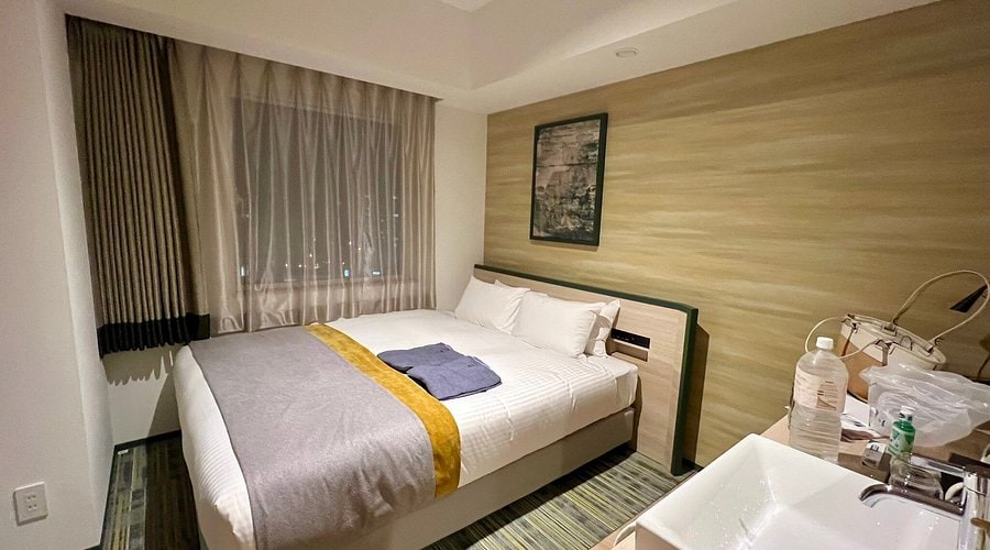 A small, modern hotel room with a double bed, gray and yellow bedding, a window with curtains, a framed picture on the wall, and a corner sink with toiletries. A folded towel lies on the bed.