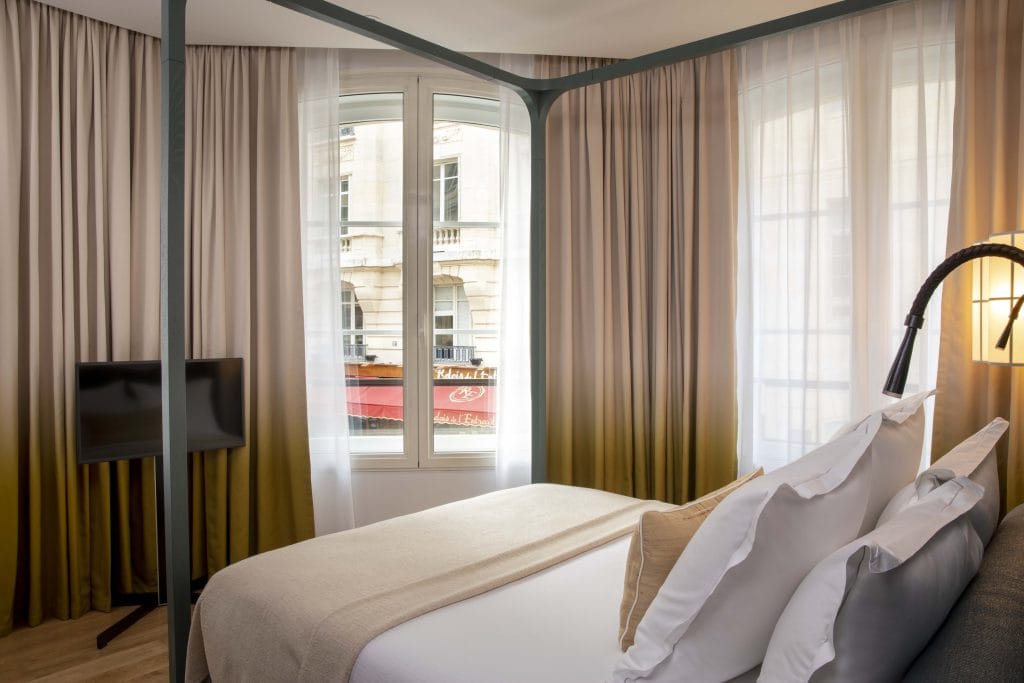 A cozy hotel room with a canopy bed, white and beige bedding, large windows with sheer curtains, a floor lamp, and a flat-screen TV on a stand. Outside, a building and red awning are visible through the window.
