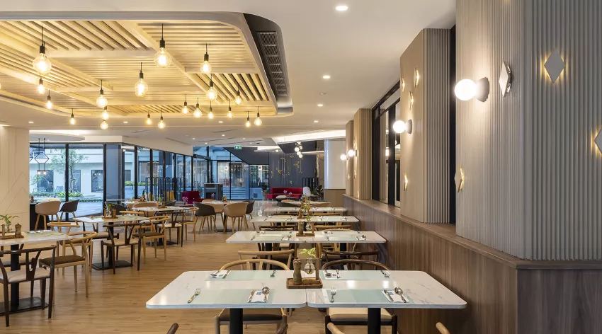 Modern restaurant interior with wooden tables and chairs, set for dining. Warm lighting from ceiling fixtures and wall sconces creates a cozy atmosphere. Large windows offer views outside, and the space is neat and inviting.