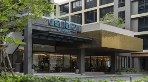 The exterior entrance of a modern hotel building with large glass windows, green plants, and a sign reading Centra by Centara above the doorway.