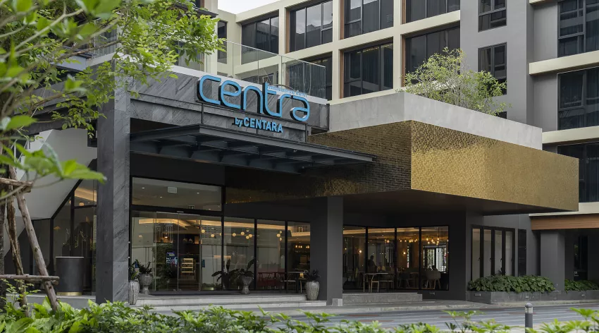 The exterior entrance of a modern hotel building with large glass windows, green plants, and a sign reading Centra by Centara above the doorway.