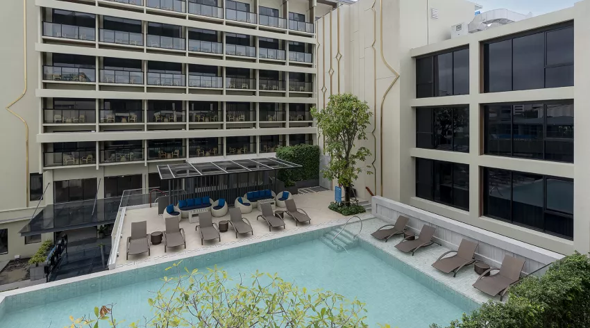 A modern hotel courtyard with a swimming pool, lounge chairs partly submerged in the water, patio seating under a canopy, and multi-story buildings surrounding the area.