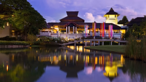 A brightly lit resort with multiple pavilions and colorful lights is reflected in a calm pond at dusk, surrounded by lush greenery and a grassy area in the foreground.