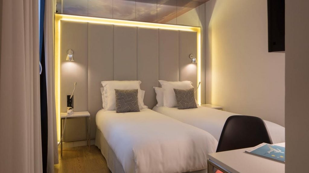 A modern hotel room with two single beds, each with white bedding and a decorative pillow, soft lighting above the headboard, two bedside tables with lamps, and a desk with a chair in the foreground.