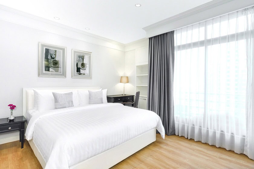 A modern, bright bedroom with a large white bed, grey and white pillows, two nightstands with lamps, framed art on the wall, a desk with a chair, and large windows with sheer curtains.