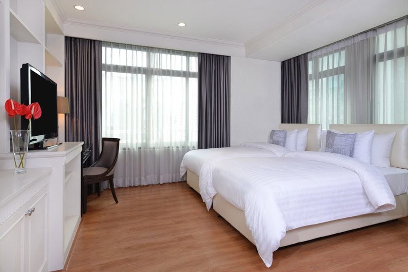 A modern hotel room with two large beds, white bedding, a desk with a chair, a flat-screen TV, and large windows with sheer and dark curtains. A vase with red flowers is on a cabinet. The floor is light wood.