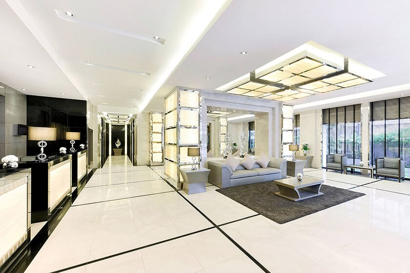 Spacious modern lobby with white tiled floors, black accents, a reception desk, a gray sofa on a dark rug, illuminated ceiling panels, and large windows with blinds letting in natural light.
