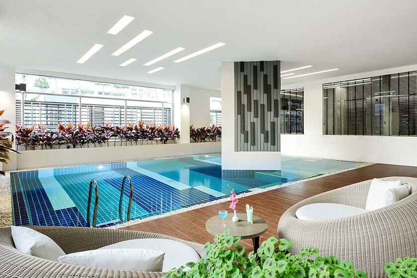 Modern indoor swimming pool with blue tiles, surrounded by lounge chairs and round cushioned seating. Large windows let in natural light and plants line the window area, creating a bright and inviting atmosphere.