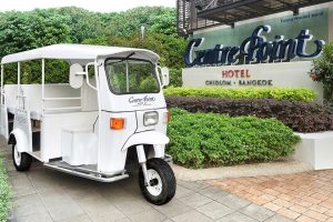 A white tuk-tuk parked on a paved path in front of the Centre Point Hotel sign, surrounded by greenery. Signs mention CHIDLOM, BANGKOK, and some Thai script.