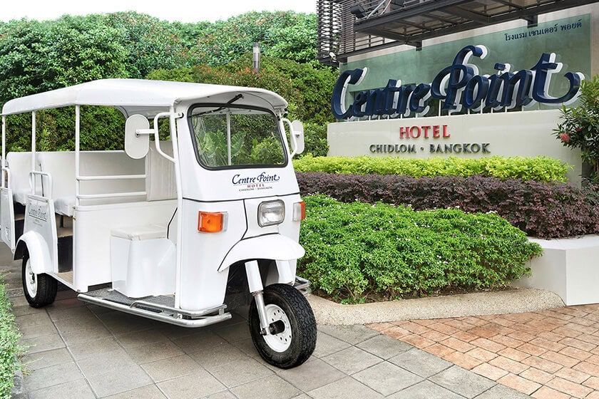 A white tuk-tuk parked on a paved path in front of the Centre Point Hotel sign, surrounded by greenery. Signs mention CHIDLOM, BANGKOK, and some Thai script.