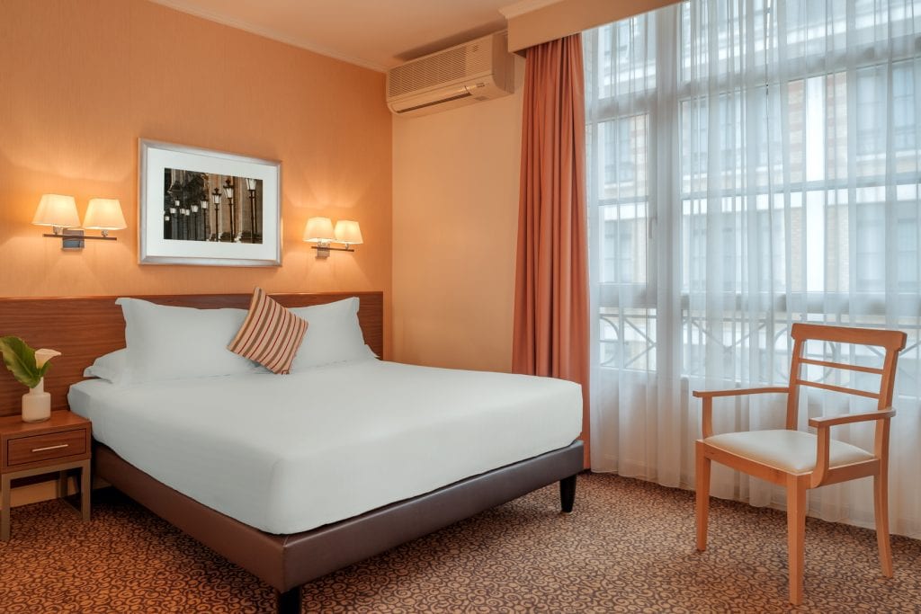 A hotel room with a double bed, white bedding, striped pillow, wall lamps, framed picture, wooden nightstand, wooden chair, carpeted floor, window with sheer curtains, and peach-colored walls and drapes.