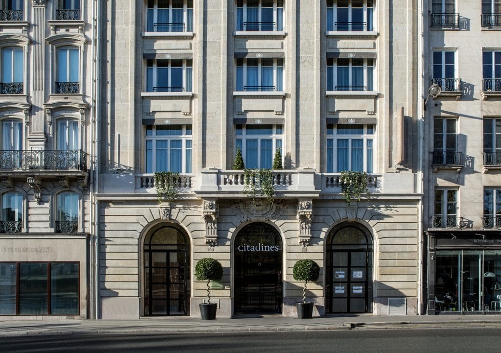 The exterior of a grand, multi-story stone building with large windows, balconies, and arched entrances. The entrance displays a citadines sign and is flanked by two round potted plants.