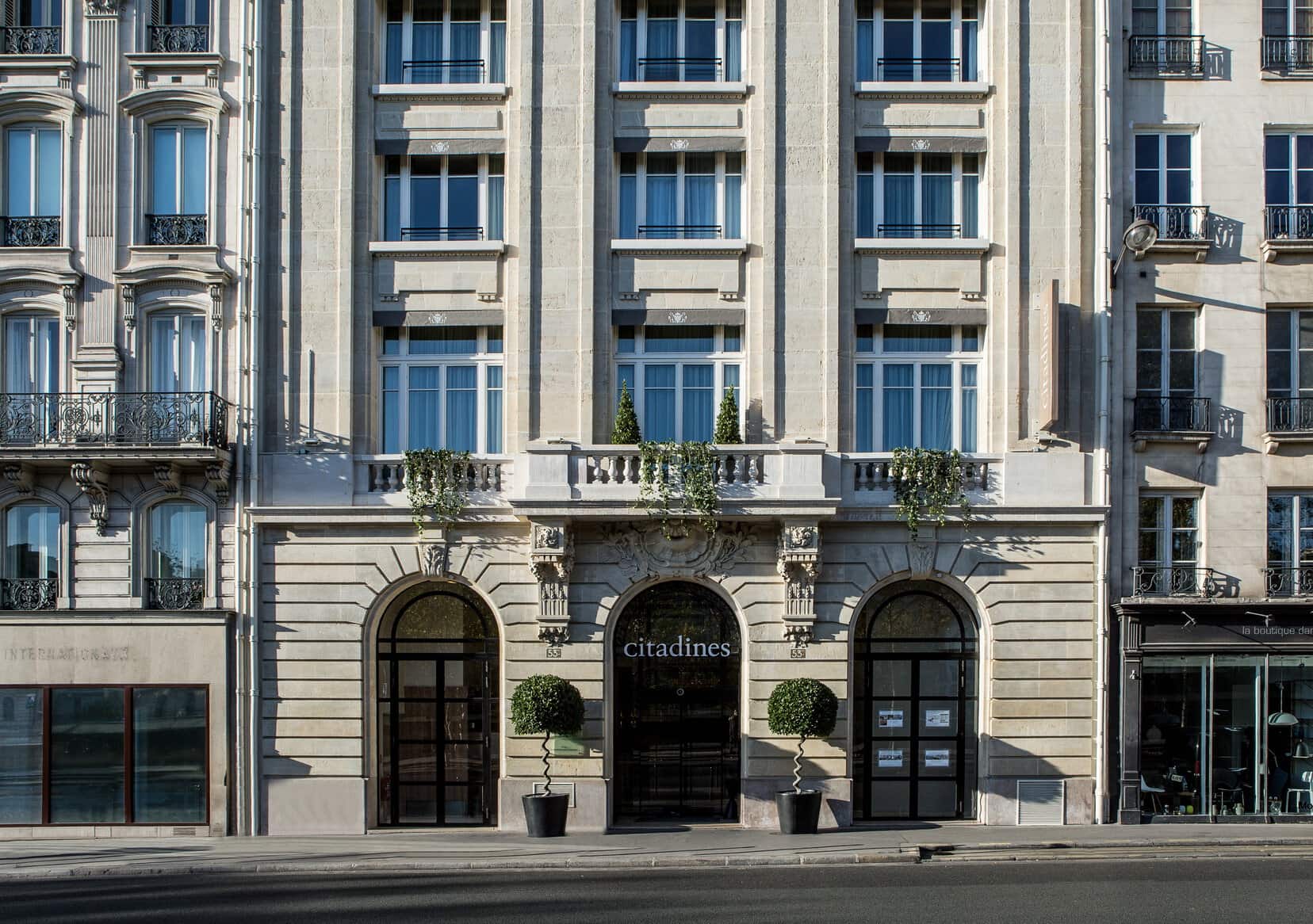 The exterior of a grand, multi-story stone building with large windows, balconies, and arched entrances. The entrance displays a citadines sign and is flanked by two round potted plants.