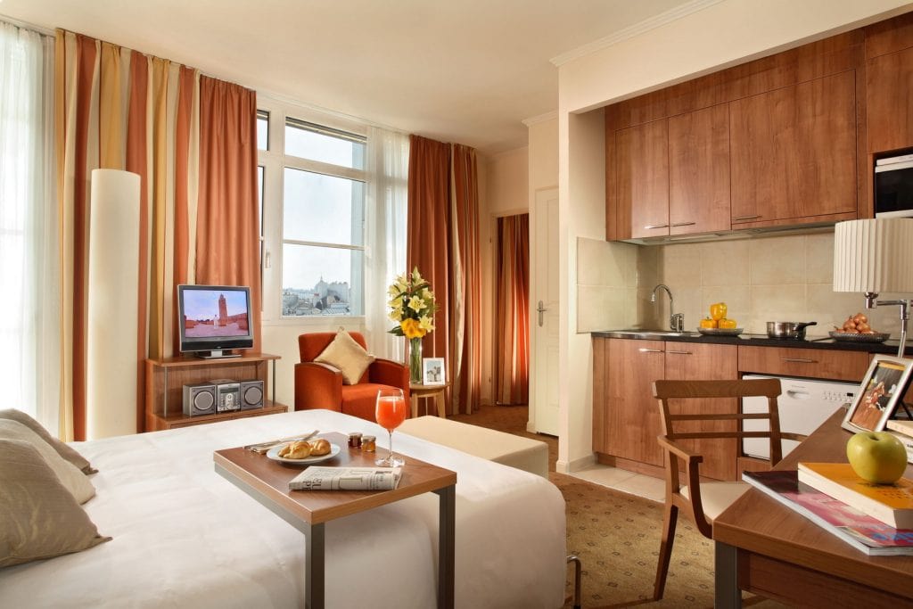 A cozy hotel room with a double bed, a small dining table set for breakfast, a kitchenette, a TV, and large windows with striped curtains letting in natural light. Fresh flowers and fruit add a welcoming touch.