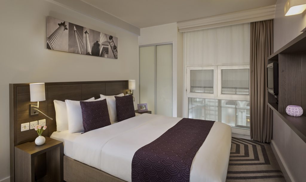 Modern hotel room with a double bed, white linens, and purple accents. Nightstands with lamps and decor sit beside the bed. Large window with sheer curtains lets in natural light. Black-and-white photo hangs above the headboard.