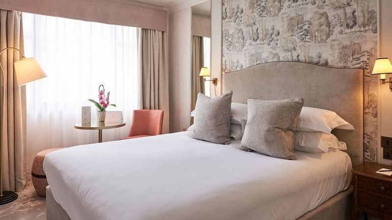 A cozy hotel room with a neatly made double bed, light gray upholstered headboard, two large pillows, bedside lamps, a round table with flowers, a pink chair, and soft natural light from a large window.