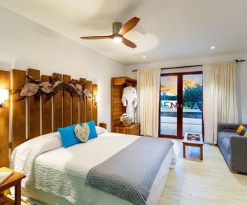 A modern, well-lit hotel room with a large bed, wood headboard, decorative pillows, a ceiling fan, a sofa, and glass doors opening to a balcony with a sunset view.