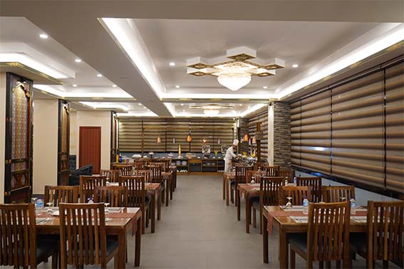 A spacious, well-lit restaurant dining area with wooden tables and chairs neatly arranged in rows. A server stands near the buffet at the back. Modern decor, large windows with brown blinds, and a chandelier overhead.