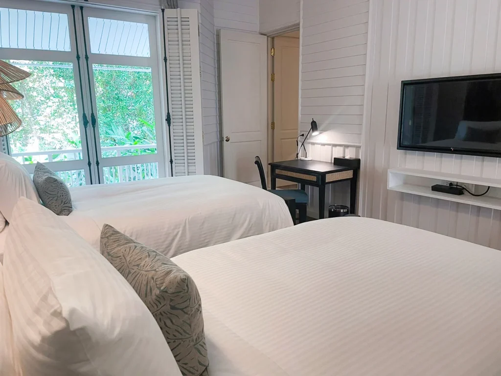 A bright hotel room with two neatly made double beds, a wall-mounted TV, a small desk with a lamp, and large windows letting in natural light with a view of greenery outside.