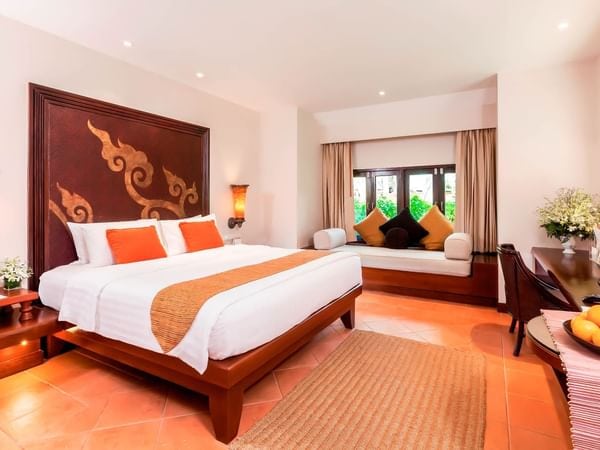 A stylish hotel room with a large bed, orange and white bedding, decorative pillows, a cushioned window seat, a rug, fresh flowers, and warm wooden furniture on terracotta tile flooring.
