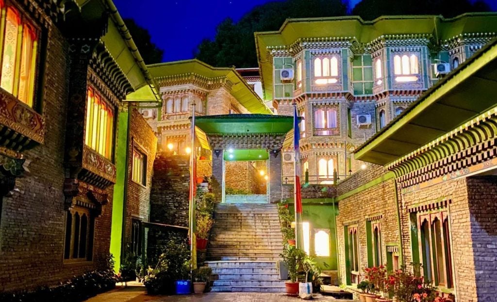 A brightly lit building with ornate windows and intricate brickwork, featuring a staircase leading to an arched entryway, surrounded by greenery at night.