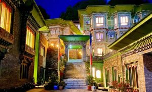 A brightly lit building with ornate windows and intricate brickwork, featuring a staircase leading to an arched entryway, surrounded by greenery at night.