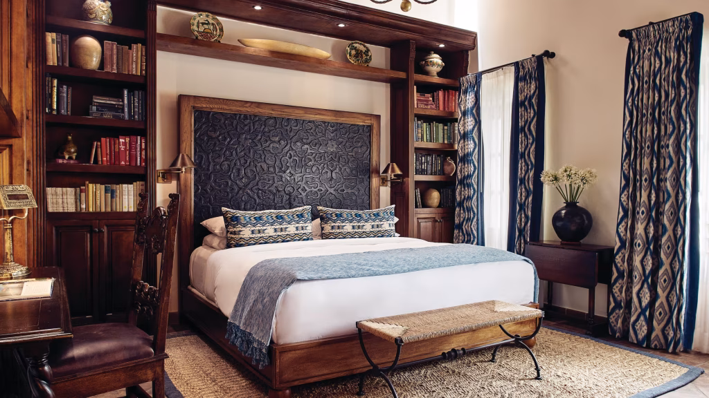 A cozy, elegant bedroom with a large wooden bed, blue-patterned headboard and bedding, bookshelves, patterned curtains, a woven bench, and decorative items on shelves and tables.