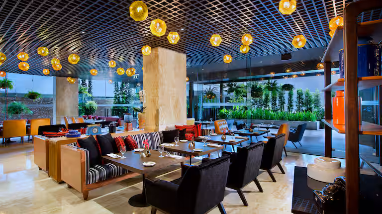 Modern restaurant interior with stylish black chairs, wooden tables, and colorful cushions; large floor-to-ceiling windows reveal lush greenery outside. Warm yellow pendant lights hang from a gridded black ceiling.