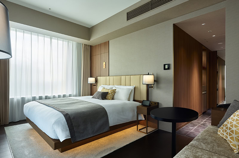 Modern hotel room with a large bed, white linens, gray throw, and accent pillows. There is a window with sheer curtains, a round black table, and a beige sofa. Warm lighting creates a cozy atmosphere.