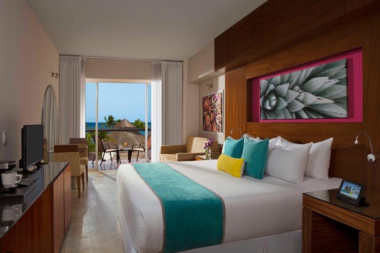 Modern hotel room with a large bed, turquoise accents, artwork above the headboard, a desk, TV, and a balcony with a view of palm trees and the ocean in the background.