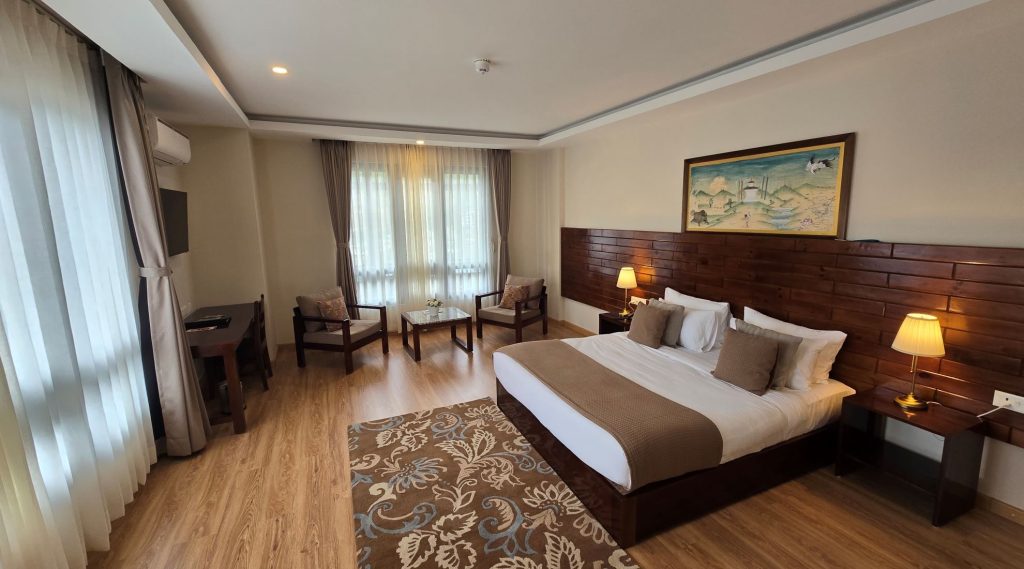 A modern hotel room with a double bed, brown and beige bedding, two armchairs near a window, a desk, a patterned rug, lamps on bedside tables, and a painting on the wall above the bed.