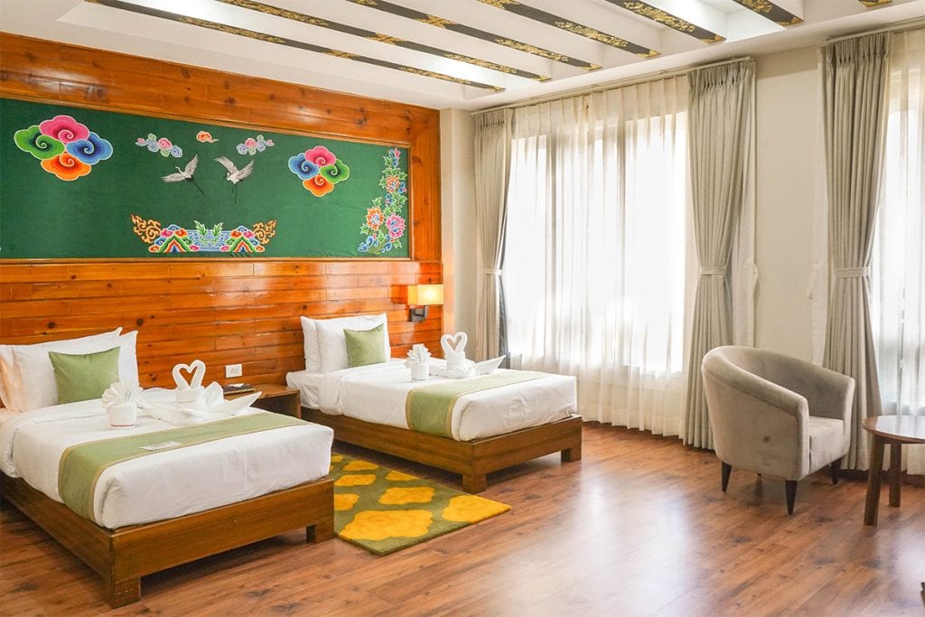 A bright hotel room with two single beds, green and white bedding, towel swans, wooden floors, a decorative mural above the beds, large windows with curtains, an armchair, and a small round table.