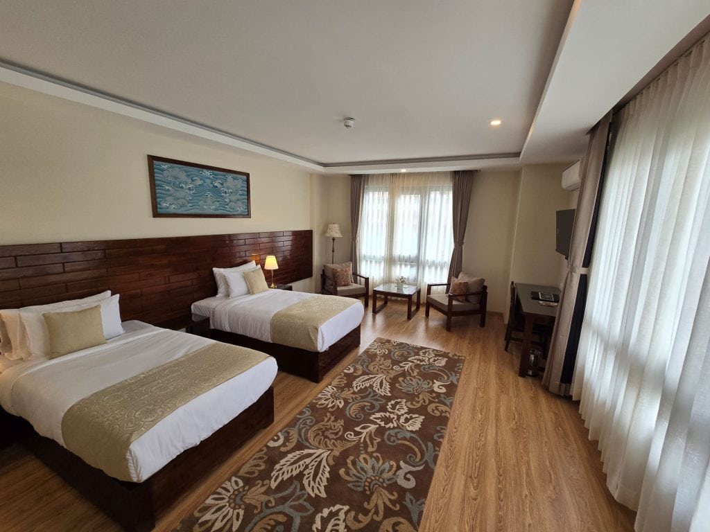 A spacious hotel room with two single beds, a wooden floor, a patterned rug, a seating area with two chairs and a small table by the window, a TV, and large windows with sheer curtains letting in natural light.