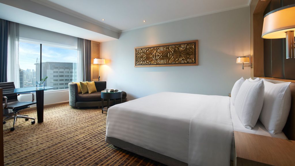 A modern hotel room with a large bed, a desk and chair by a window with city views, a cozy armchair with a small table, neutral-colored walls, and elegant decor.