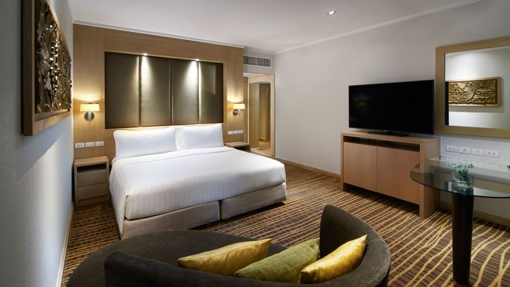 Modern hotel room with a large bed, flat-screen TV on a wooden stand, glass table, curved sofa with yellow cushions, bedside tables, and decorative wall art, all in neutral and gold tones.