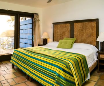 A cozy bedroom with a large bed featuring a green and yellow striped blanket, two green pillows, wicker headboard, bedside tables with lamps, tiled floor, and glass doors opening to a patio.