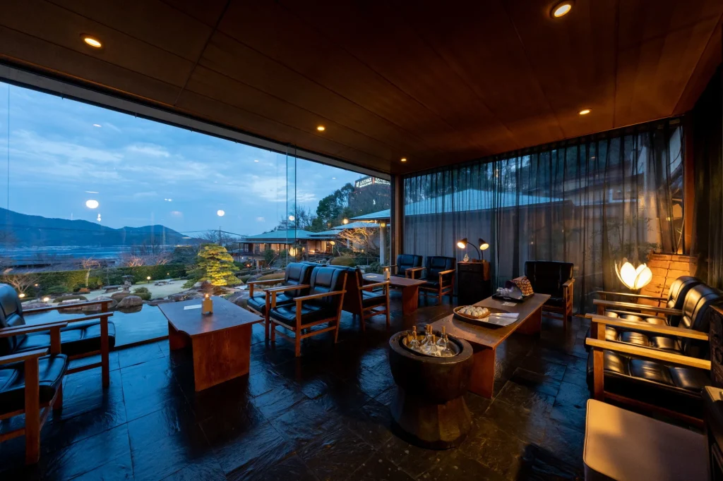 A modern lounge with dark wooden furniture and large floor-to-ceiling windows overlooking a tranquil outdoor garden and mountains at dusk. Warm lighting creates a cozy atmosphere inside.