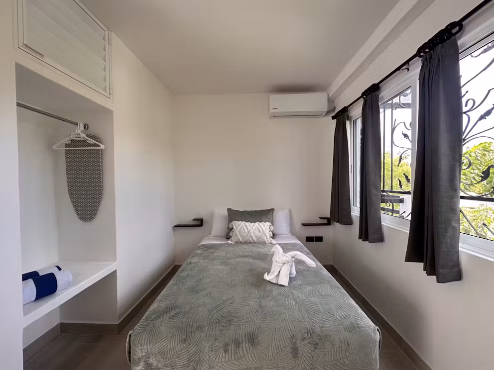 A small, minimalist bedroom with a neatly made bed, gray bedding, two pillows, towel art, large windows with dark curtains, an ironing board, and an air conditioner on the wall.