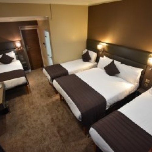 Hotel room with four single beds, each neatly made with white sheets, brown throws, and matching cushions. The room has carpeted floors, wall-mounted lights above each bed, and a closed door visible at the end.