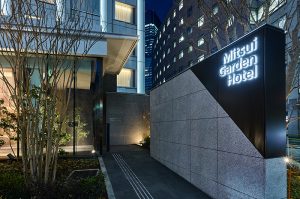 Entrance to Mitsui Garden Hotel at night, with modern buildings, illuminated sign, trees, and city lights in the background.